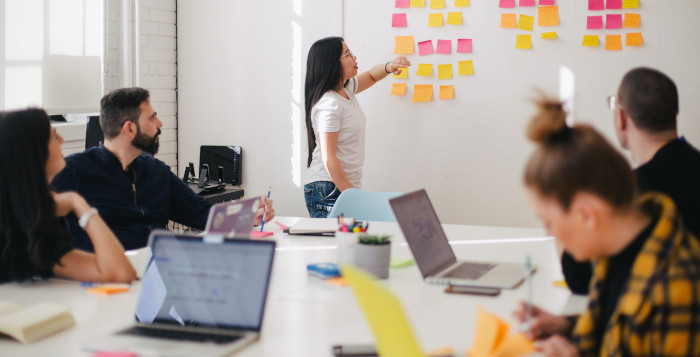 Picture of a female instructor at a whiteboard covered in post-its teaching a Health and Safety course to students sitting around a table with laptops.